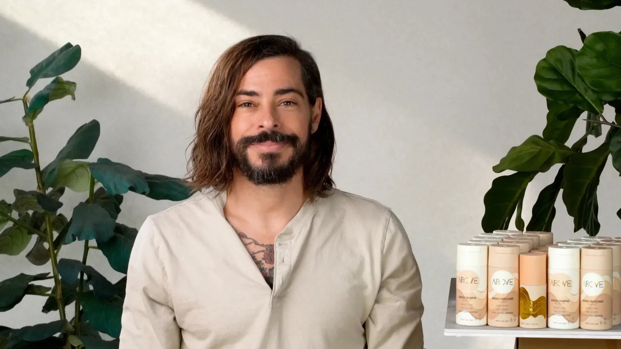 Smiling man with healthy shoulder-length hair and beard standing beside Arove Beauty waterless shampoo and conditioner display, showcasing clean sustainable haircare in a natural setting with plants.