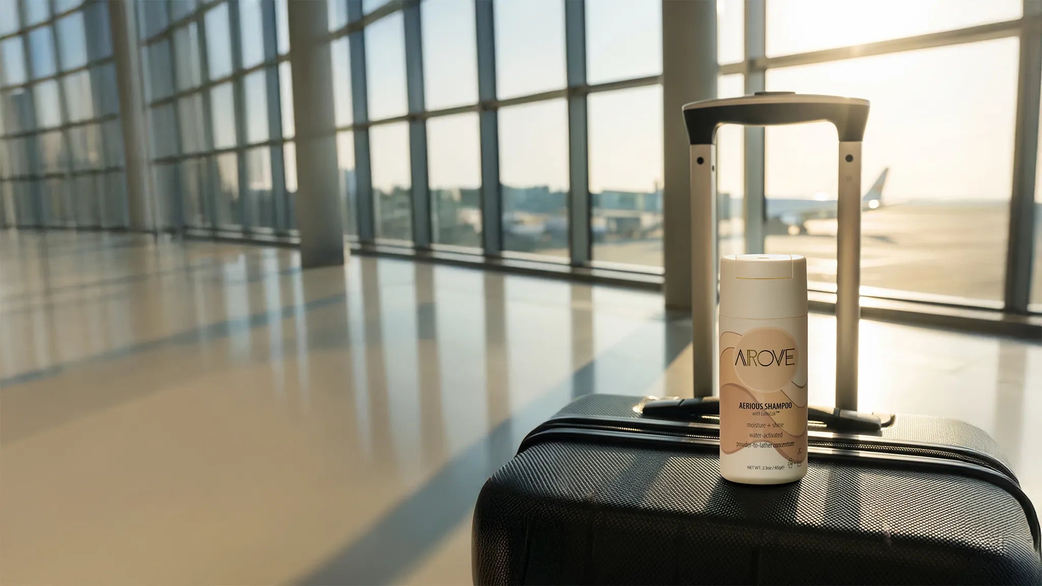 Arove Aerious Waterless Powder Shampoo sitting on a suitcase in an airport terminal, travel-friendly zero-water concentrated formula.